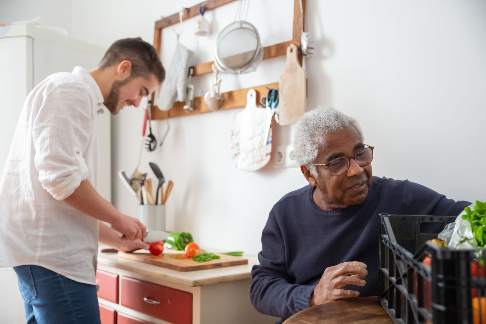 A young man chopping vegetables in a kitchen while an older neighbour sits nearby smiling