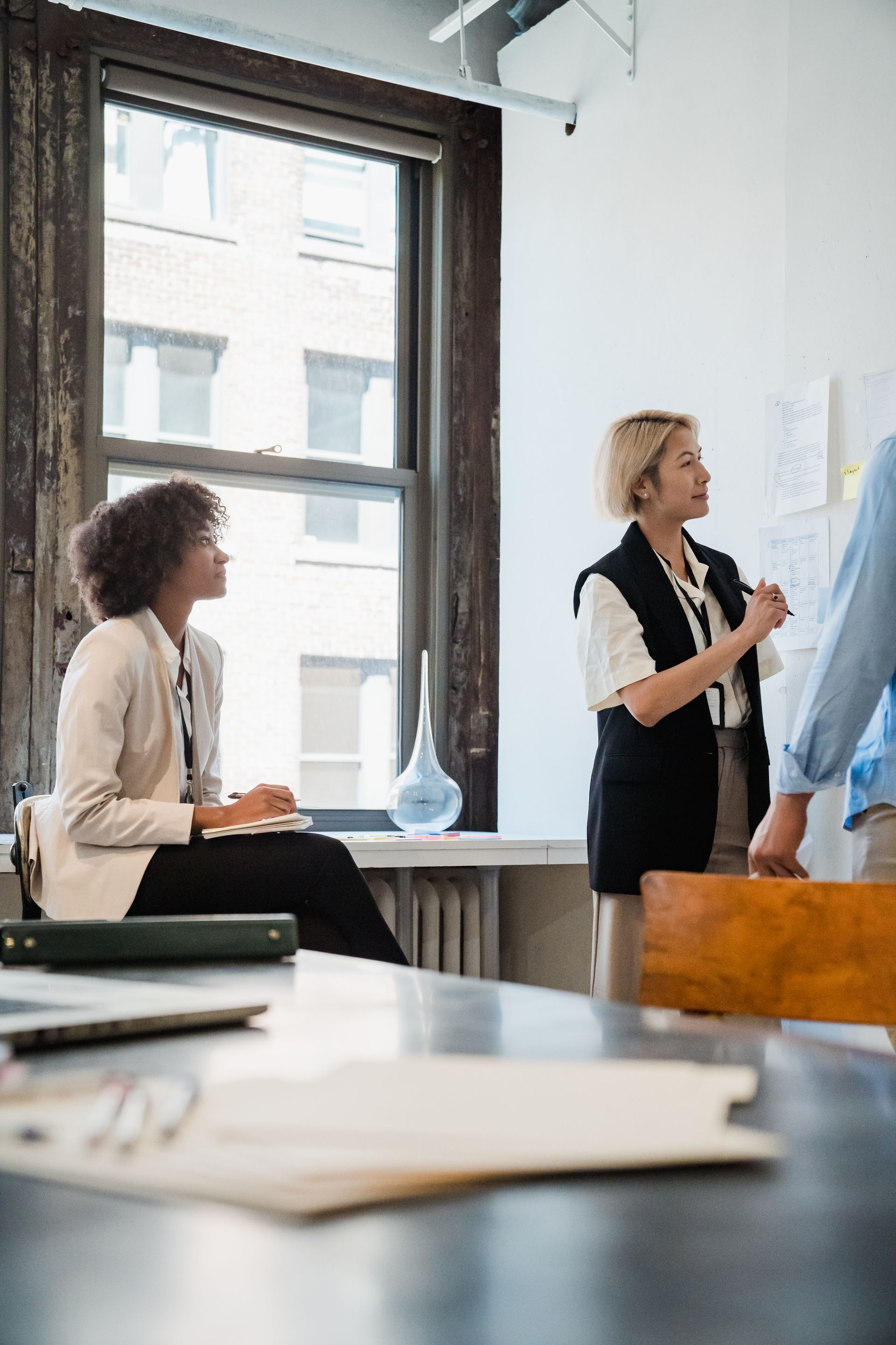 Two colleagues in a bright office with big windows, listening and taking notes