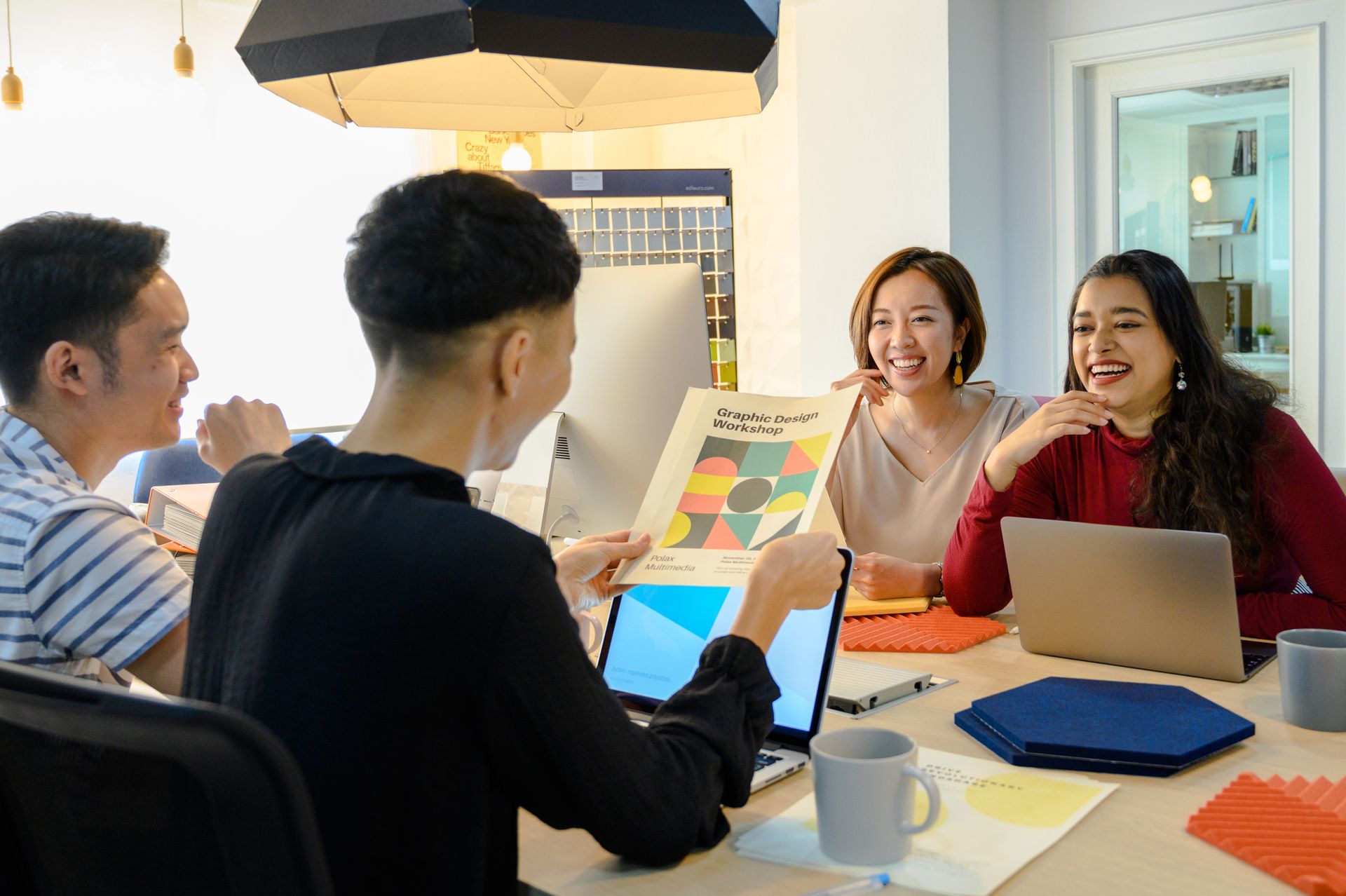A small team laughing around a table while one person holds up a printed graphic design sample
