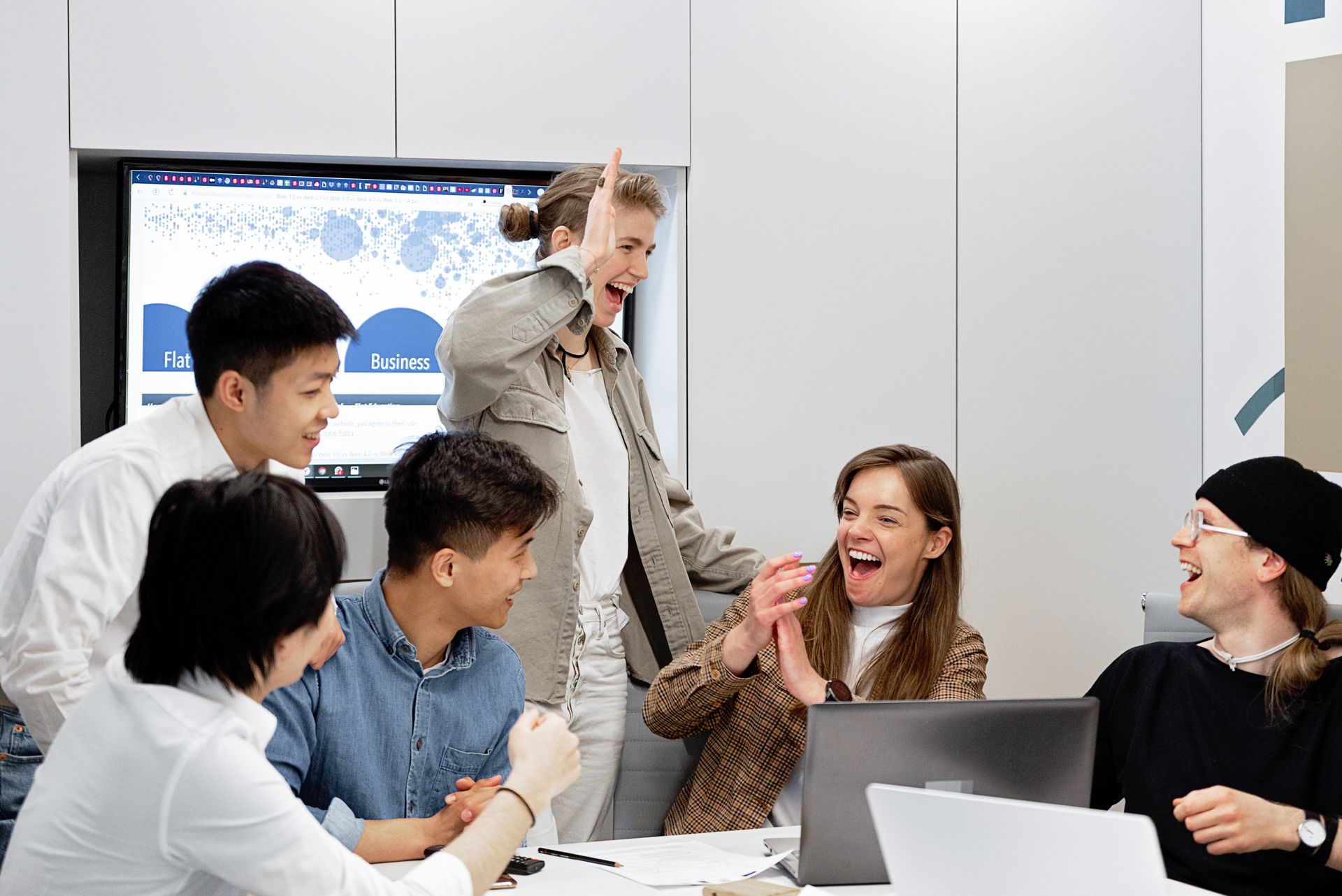 A team of colleagues laughing together around a conference table with laptops and a screen behind them