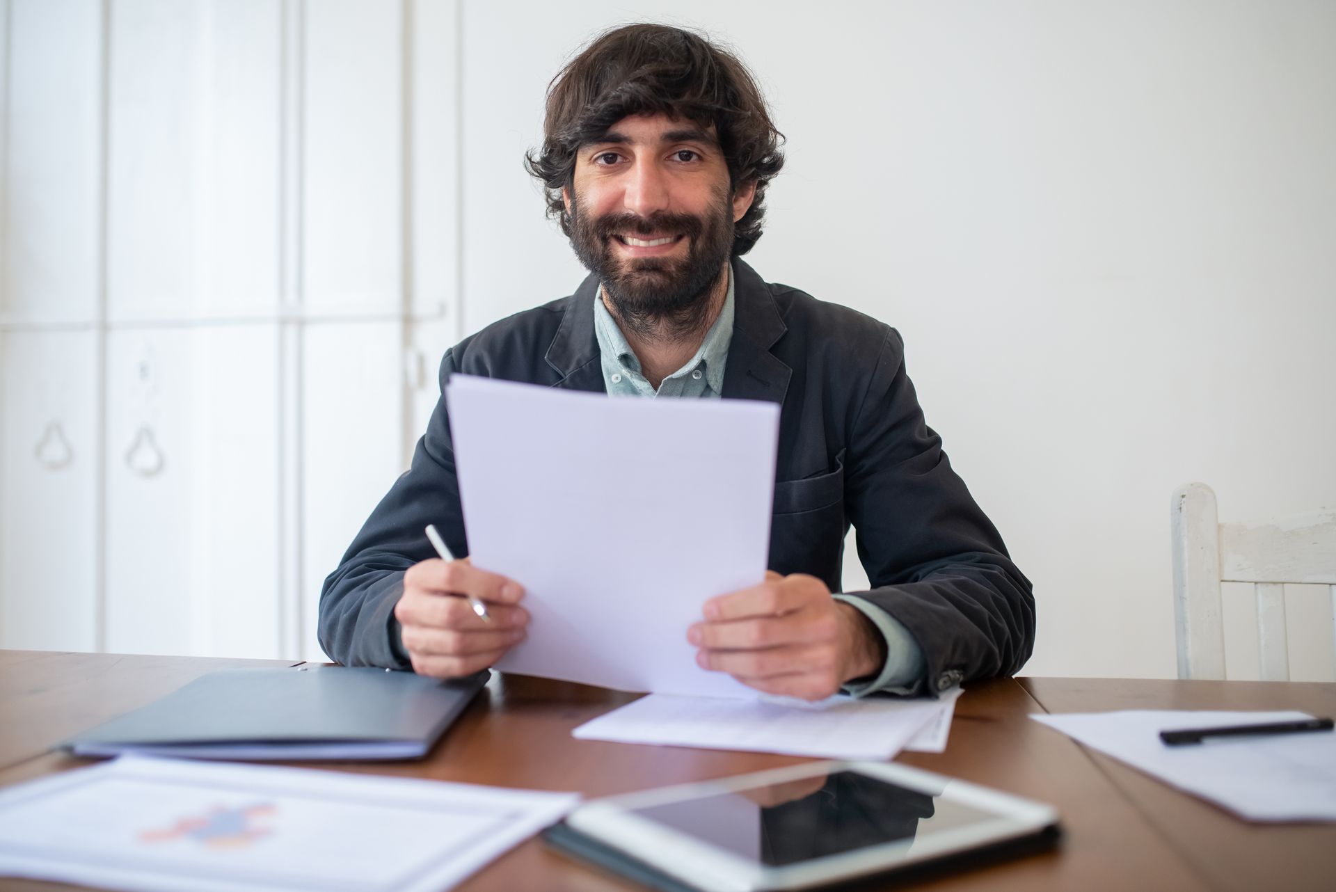 A smiling bearded man in a dark jacket holding a sheet of paper at a wooden desk