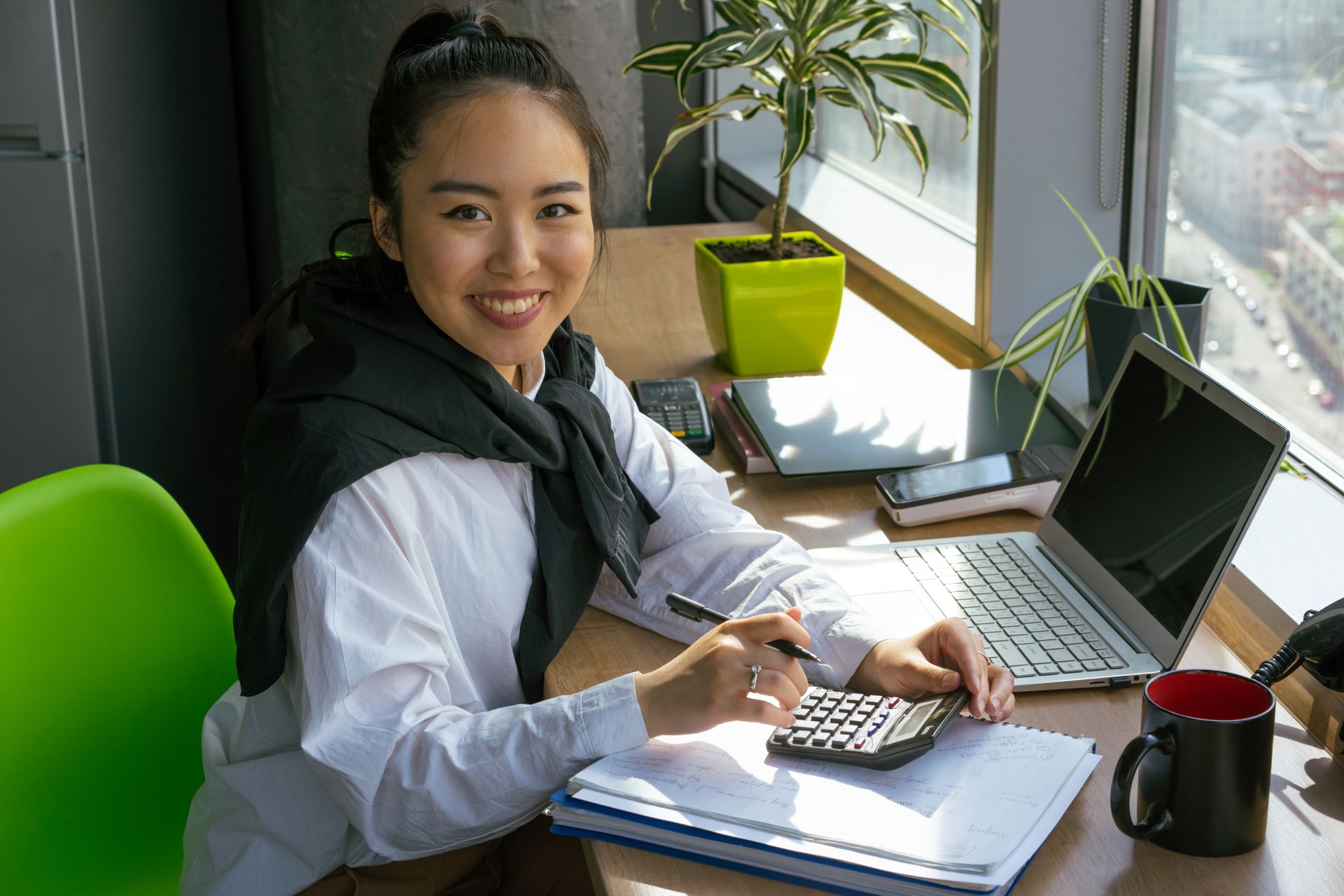 A smiling woman at a sunny window desk using a calculator beside a laptop and notebook