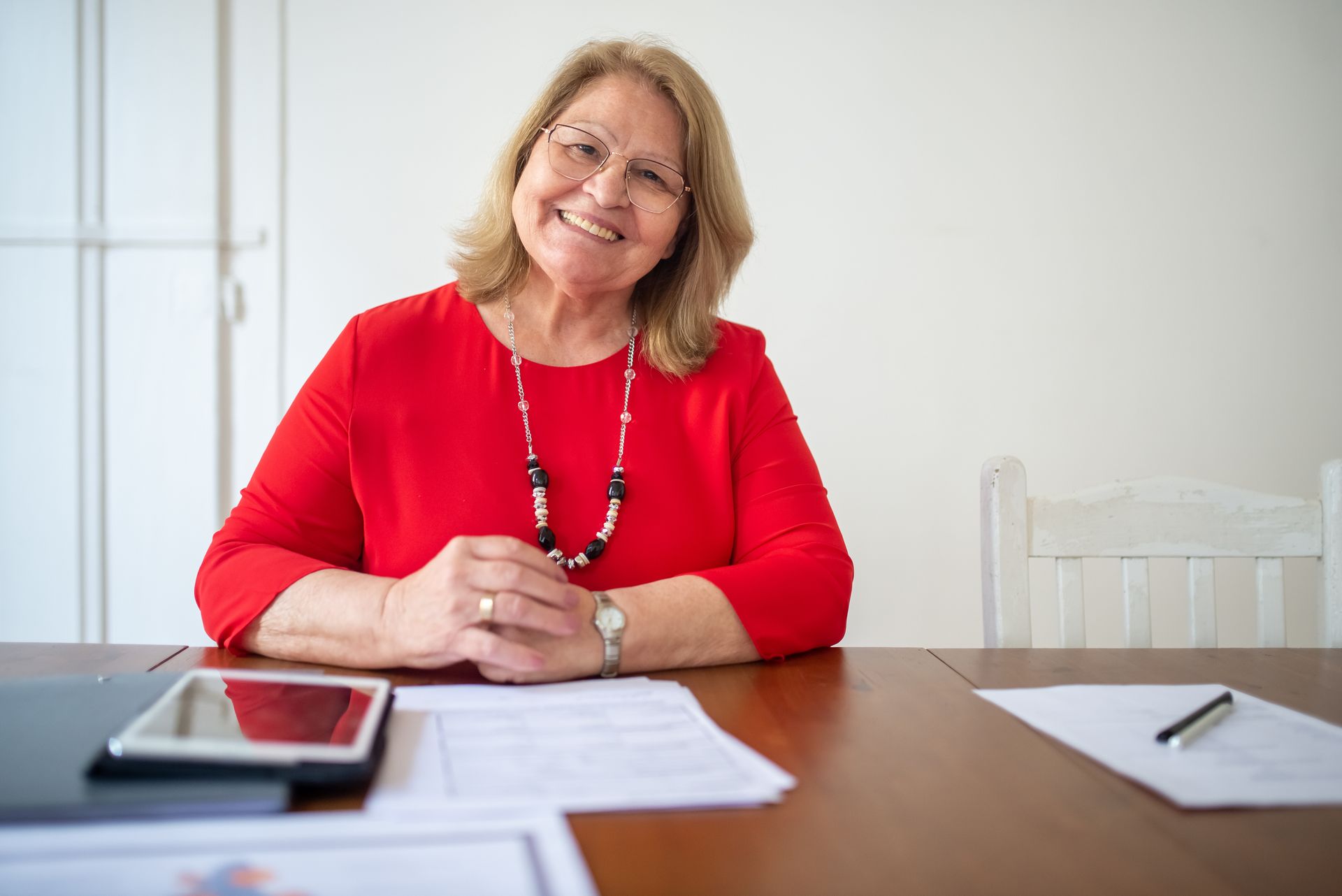 A smiling older woman in a red top with glasses sitting at a wooden table with a tablet and papers