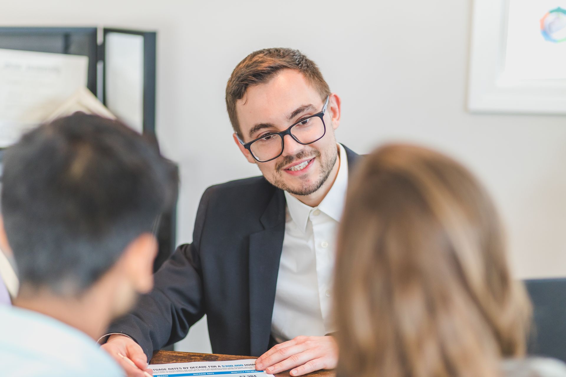 A smiling advisor in glasses and a dark blazer talking with two clients across a desk