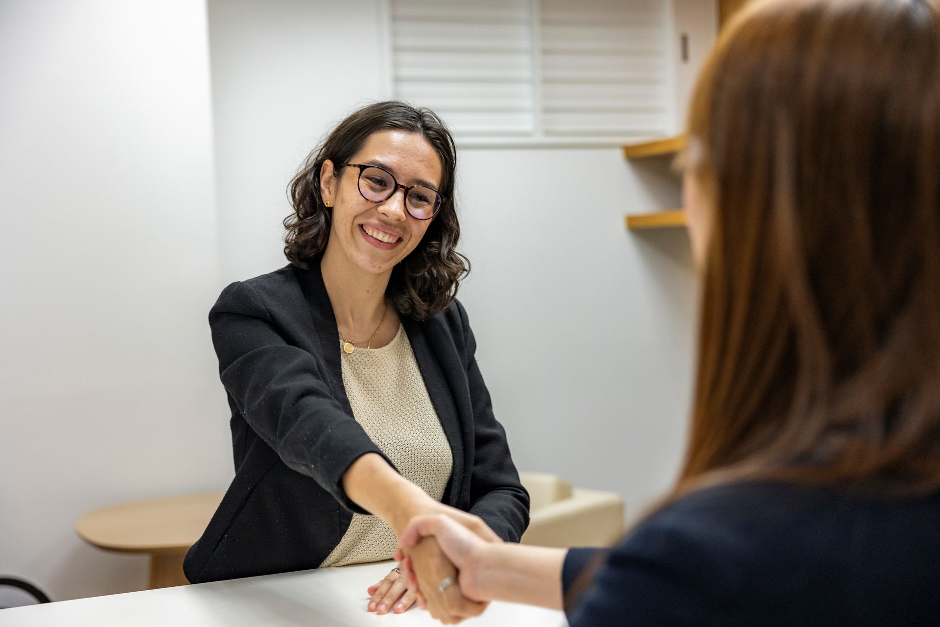 A smiling woman in a black blazer and round glasses shaking hands with a client across a white table