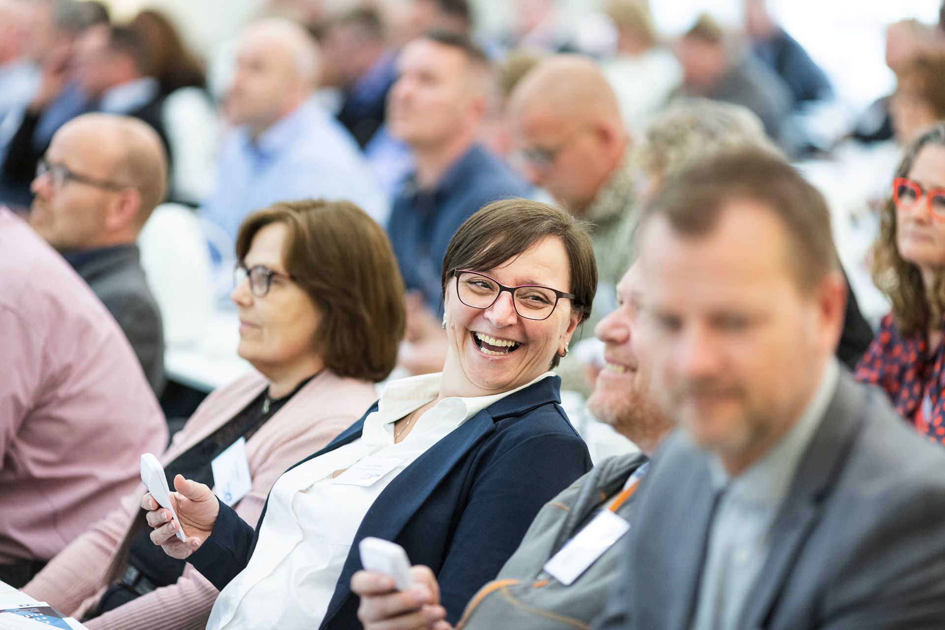 A woman in glasses laughing as she turns to a neighbour in a busy conference audience