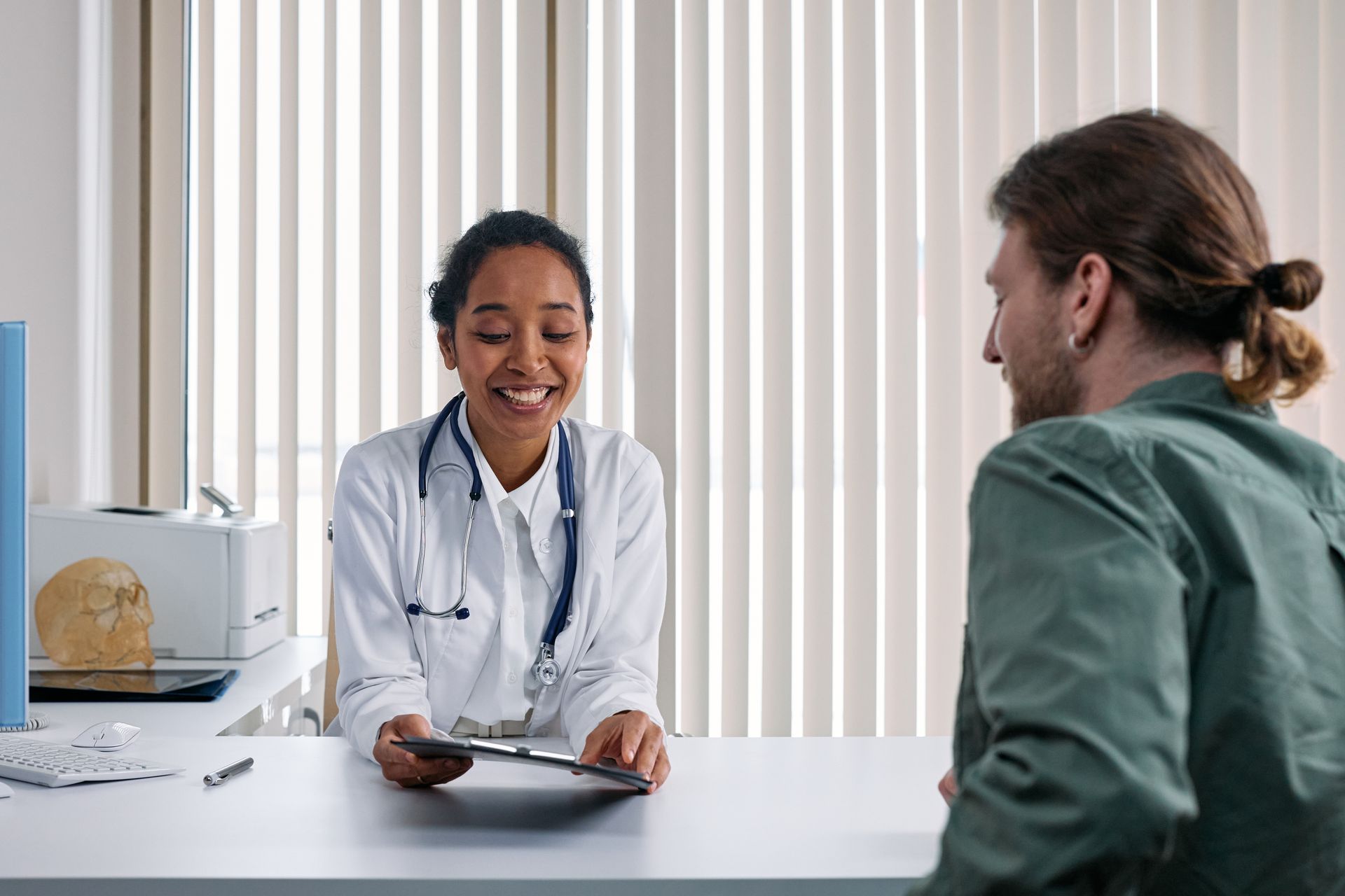 A smiling doctor at a desk showing a tablet to a patient in a bright consulting room