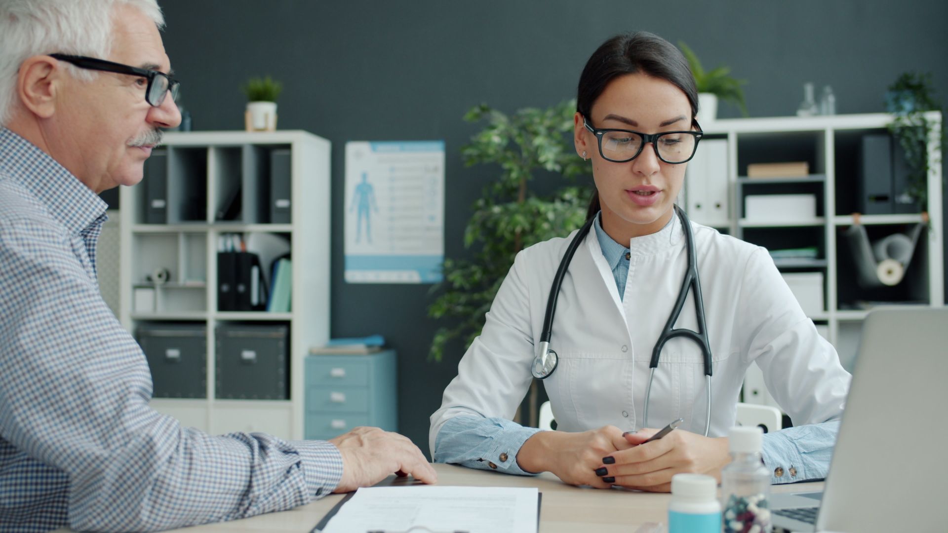 A doctor in glasses with a stethoscope listening attentively to an older patient at her desk