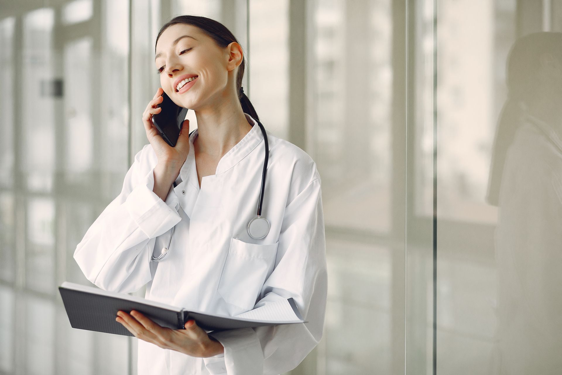 A smiling doctor in a white coat taking a phone call, holding a notebook