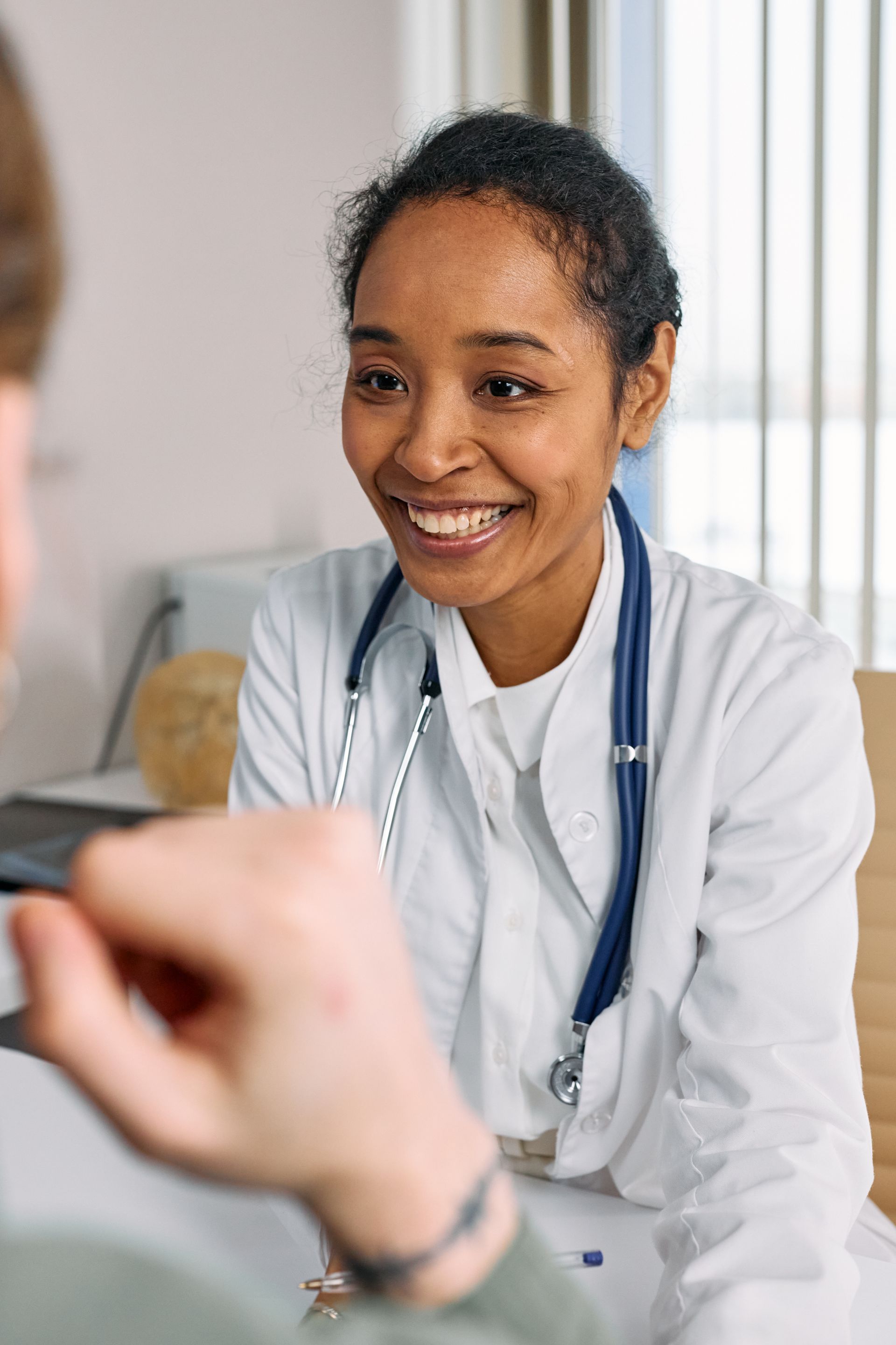 A smiling doctor in a white coat with a stethoscope listening to a patient in a bright consulting room