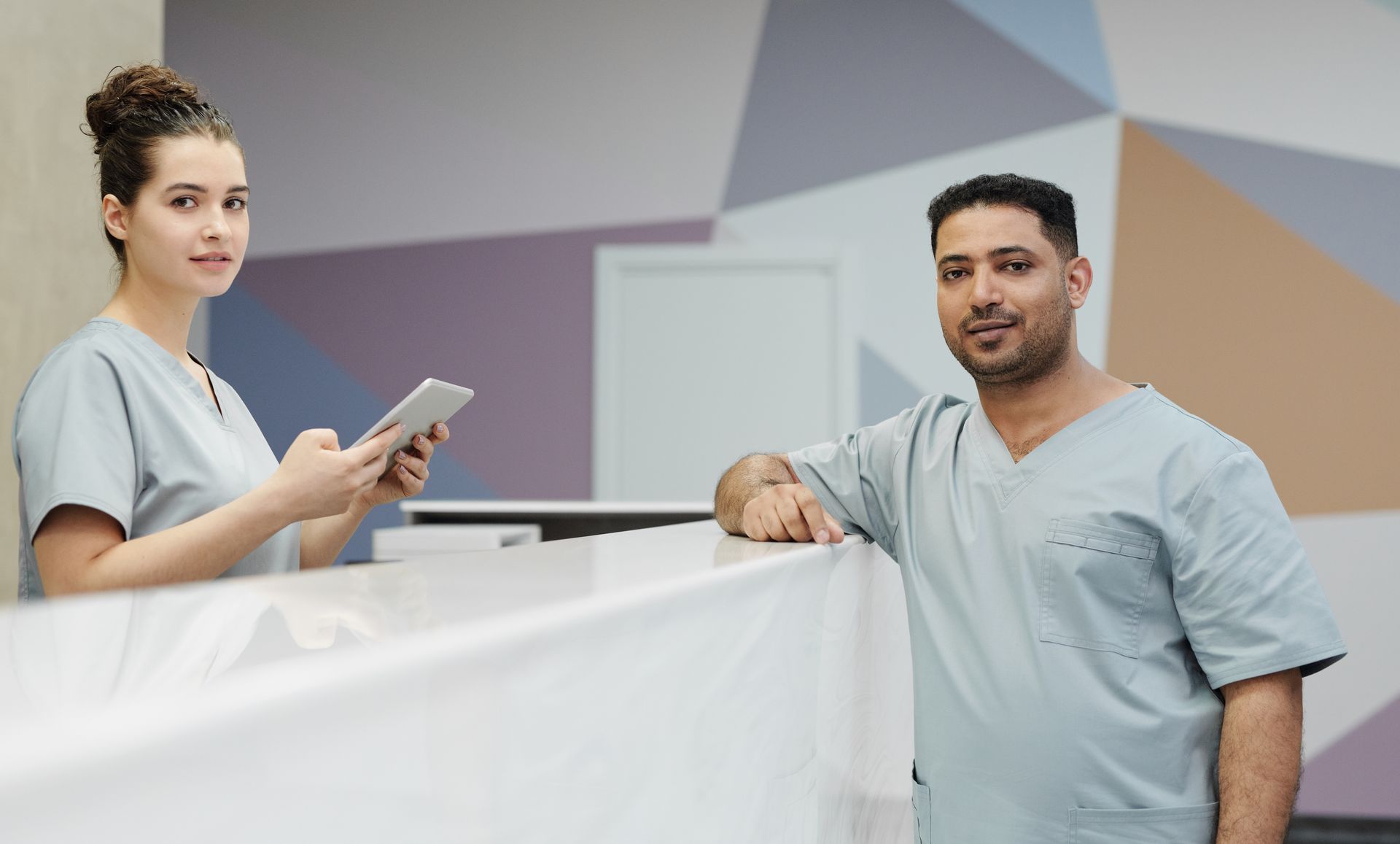 Two clinic team members in grey scrubs at a reception desk, one holding a tablet