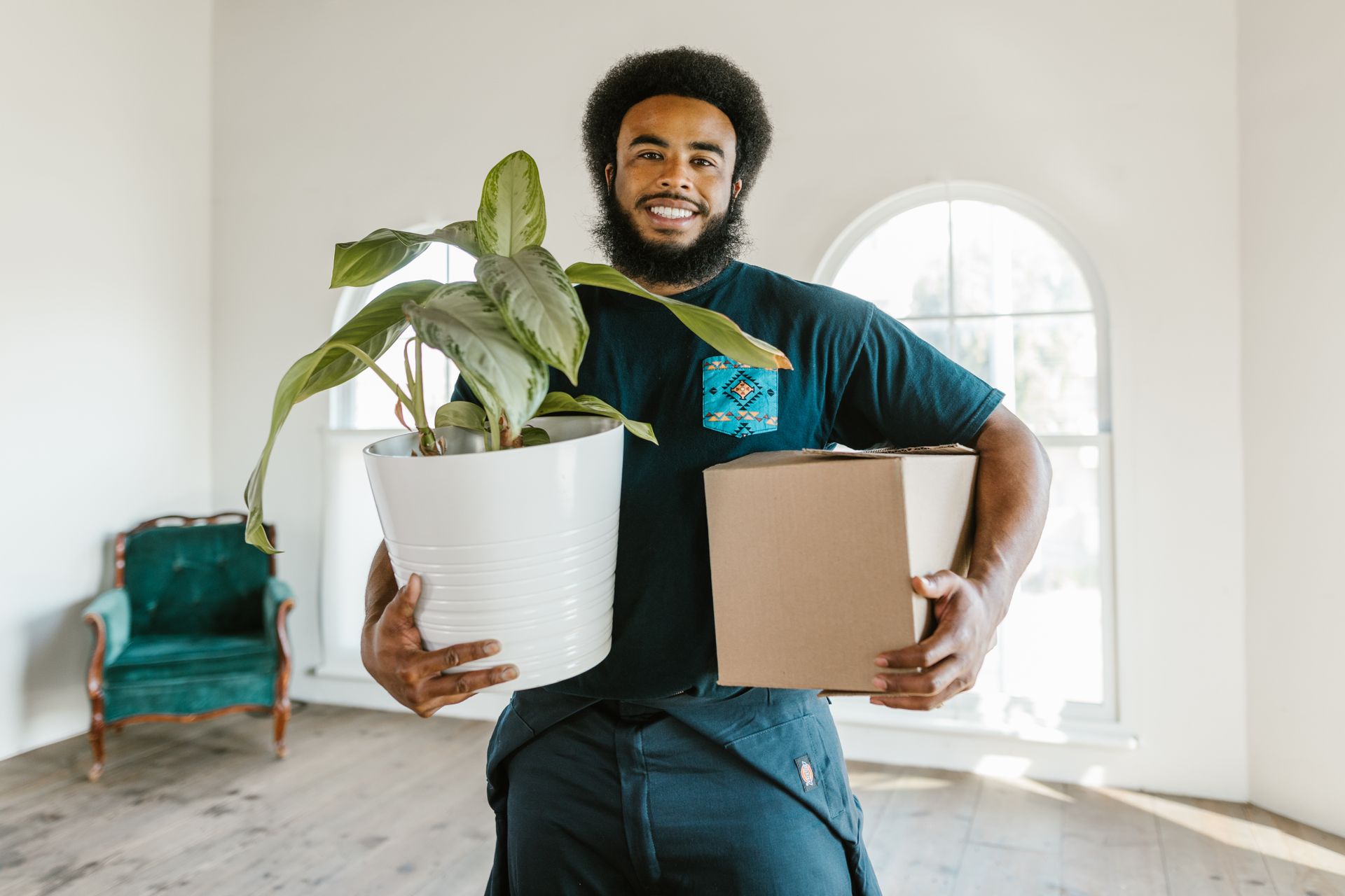 A smiling man in a dark uniform holding a potted plant and a cardboard box in a bright empty room