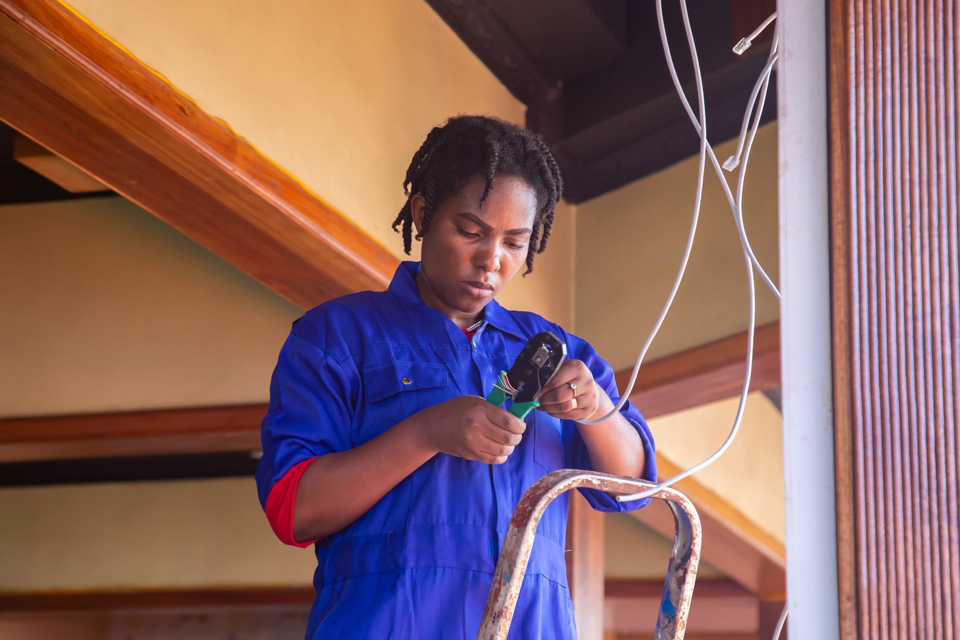 A woman in blue overalls on a ladder crimping a cable connector