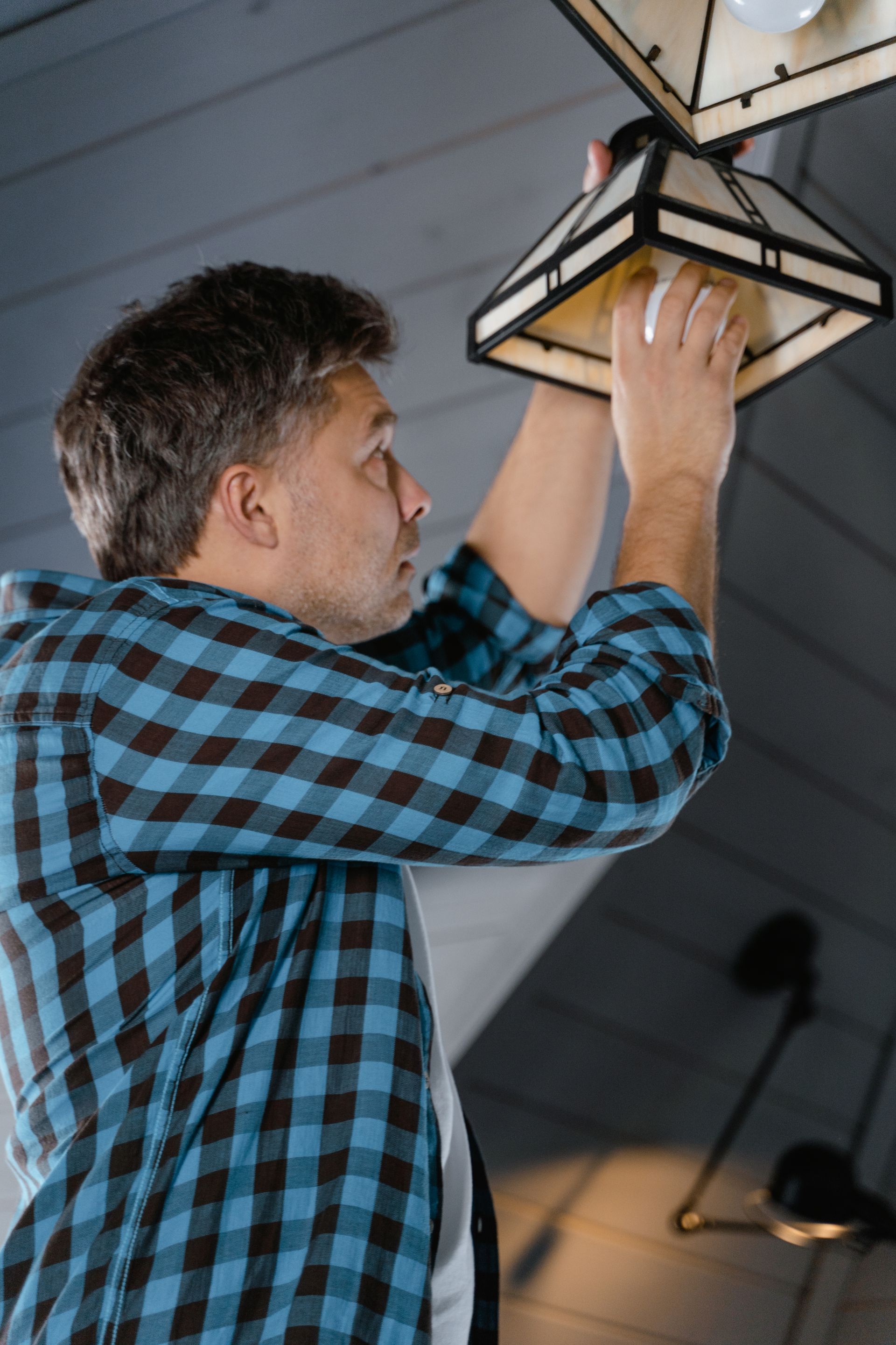 A man in a checked shirt reaching up to fit a bulb in a ceiling lantern