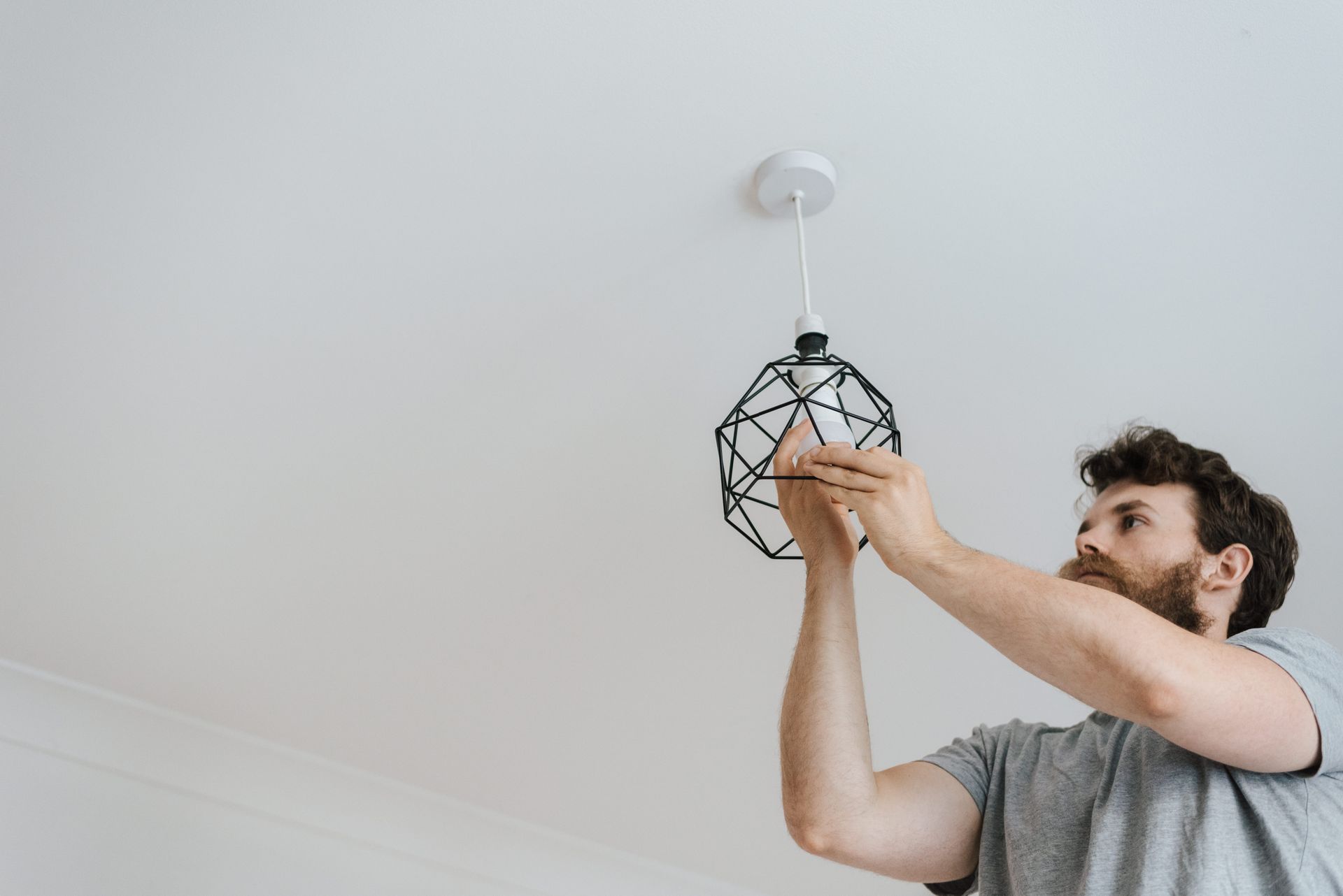 A tradesperson in a grey T-shirt fitting a light fixture to a ceiling in a bright room