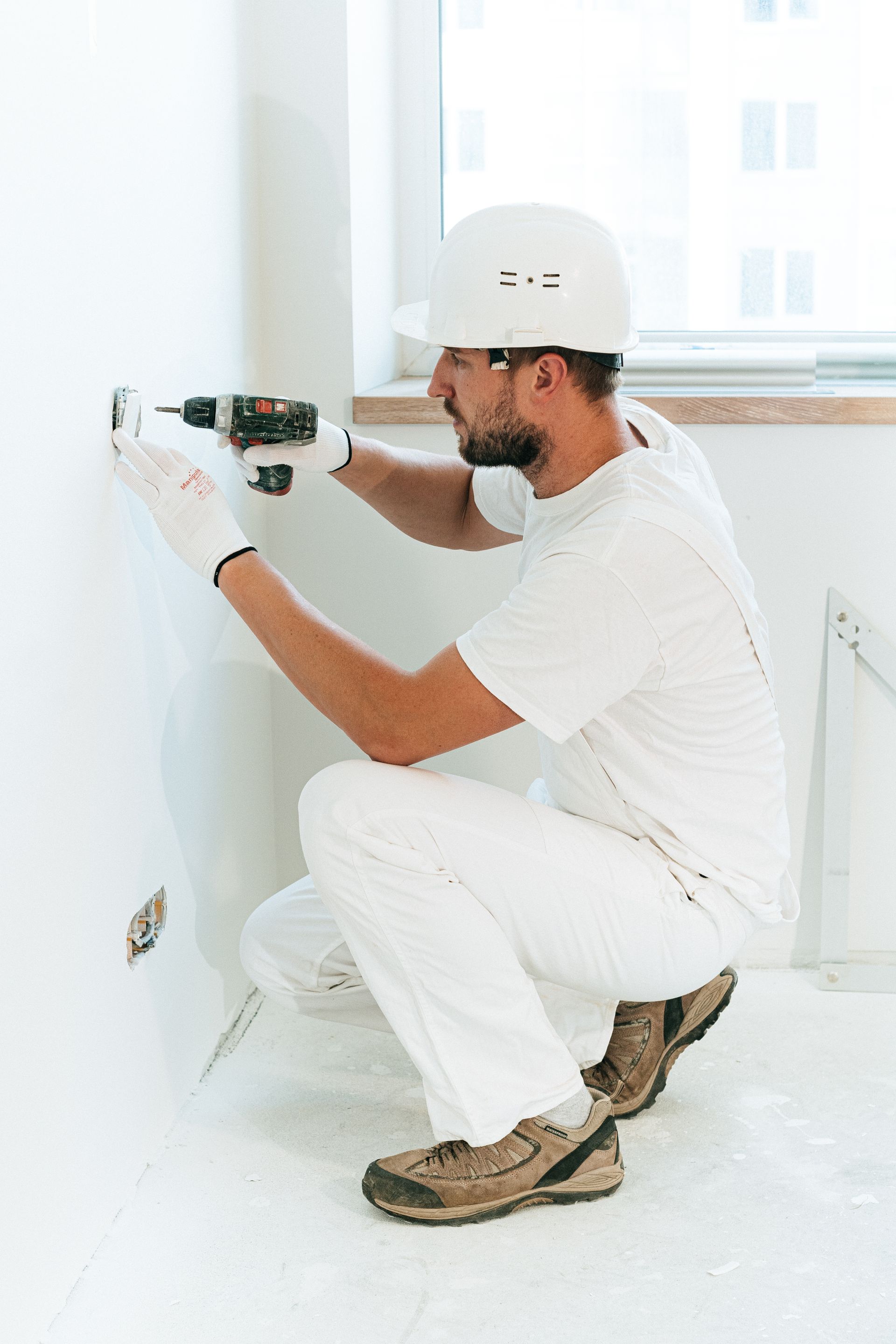 A tradesman in white overalls and a white hard hat crouching to fit a wall socket with a cordless drill