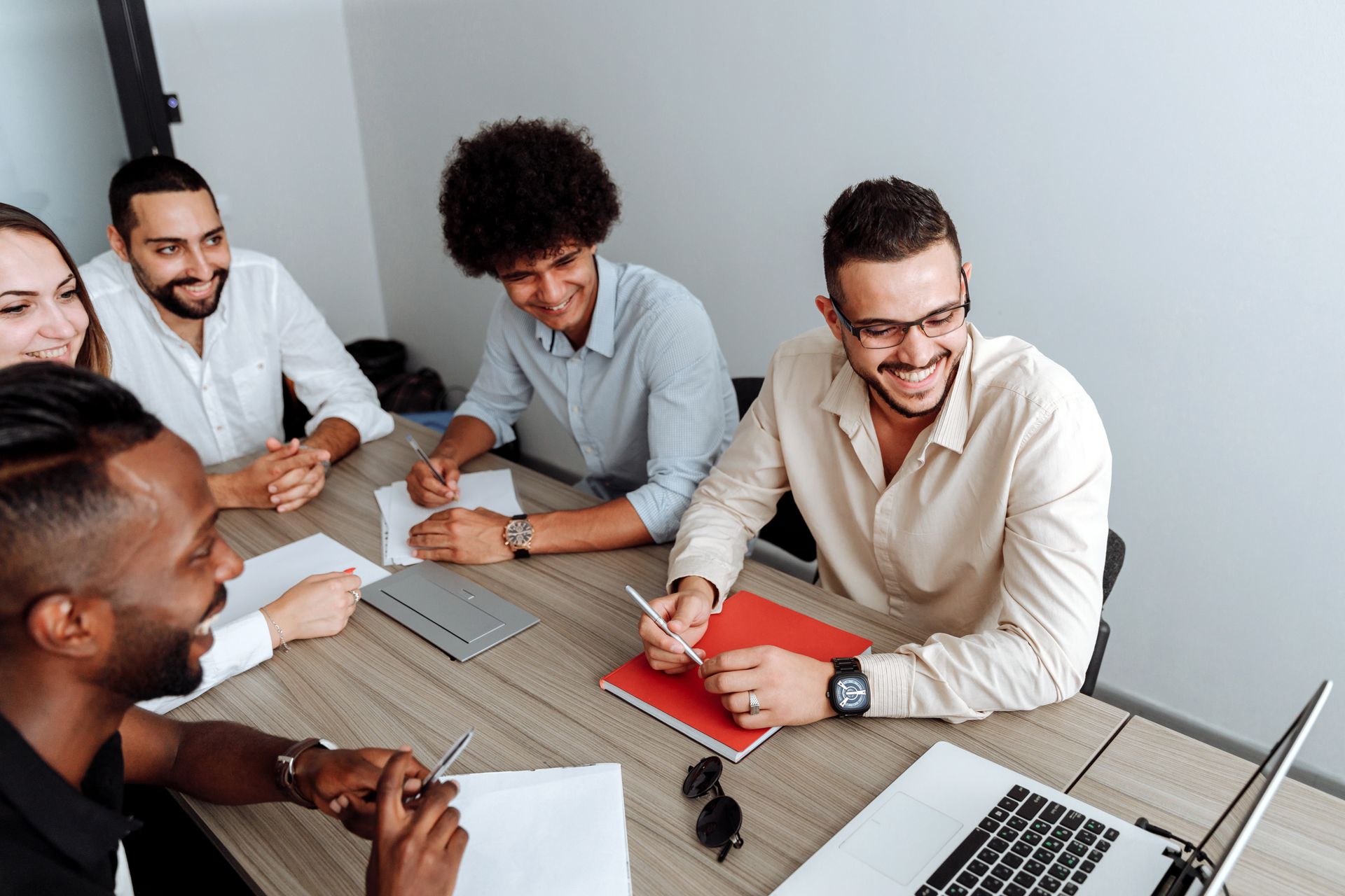 Five colleagues smiling and listening around a meeting table with notebooks and a laptop