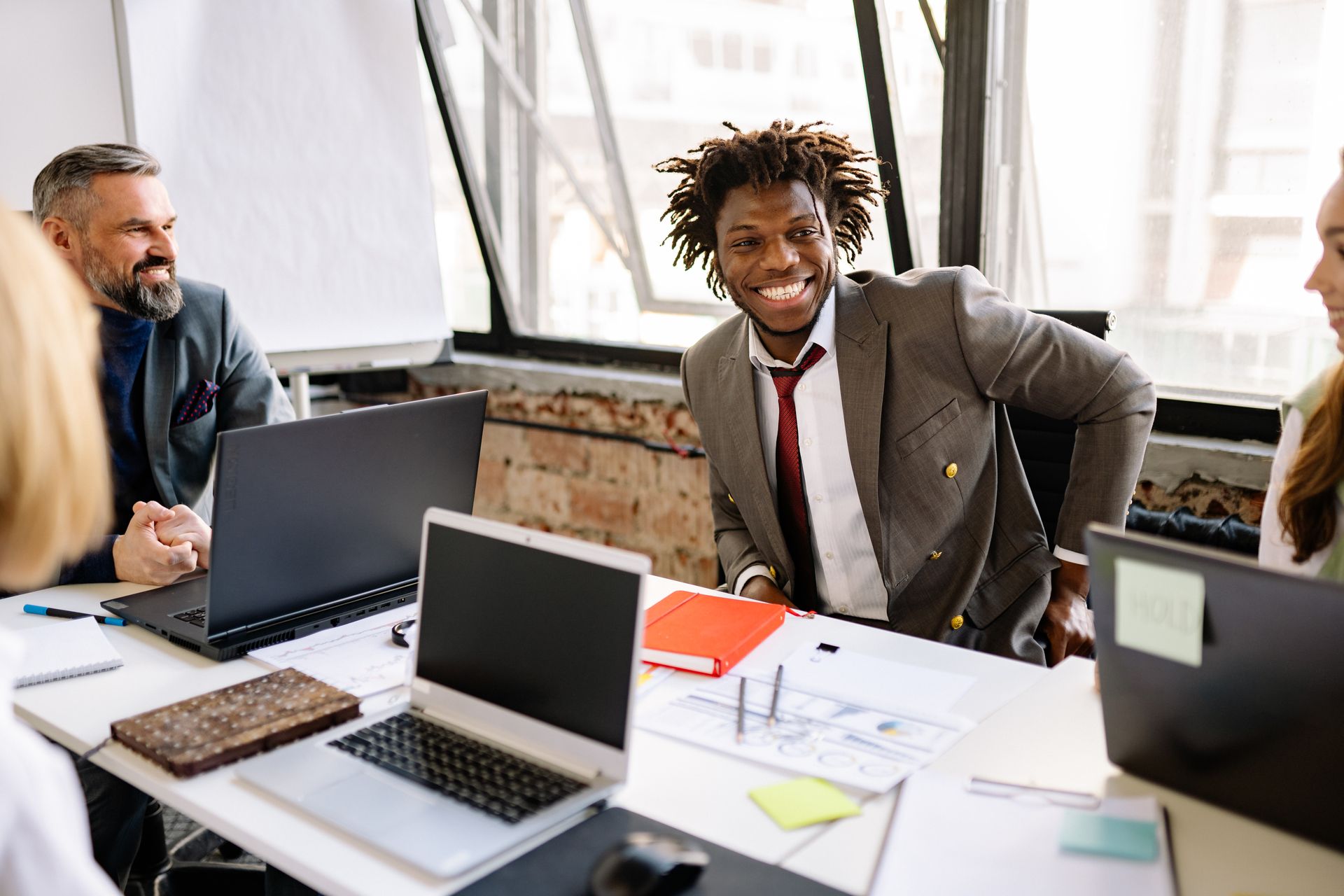 A man in a grey suit smiling warmly across a meeting table with laptops