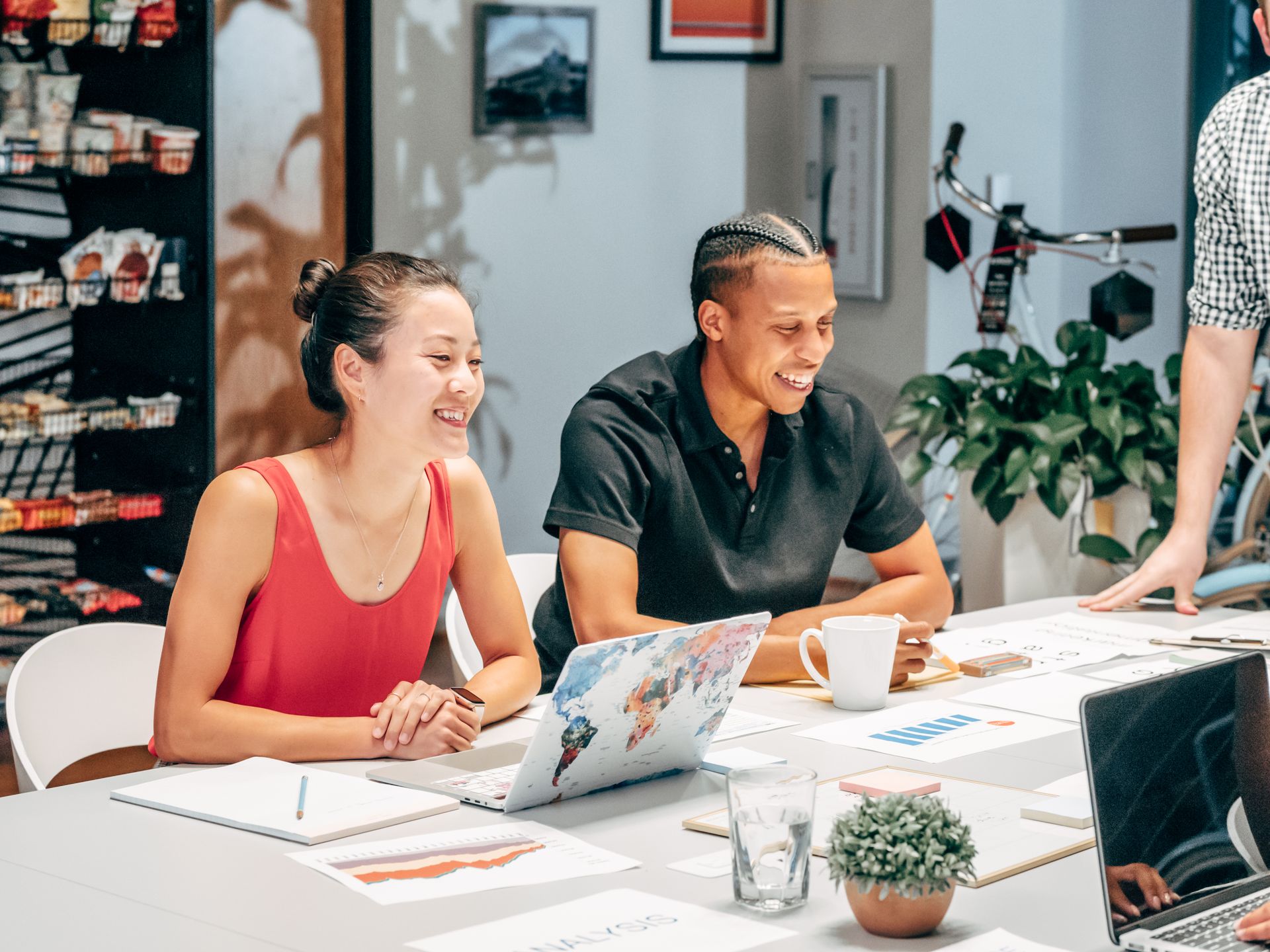 Two colleagues laughing together at a meeting table with a laptop and printed charts