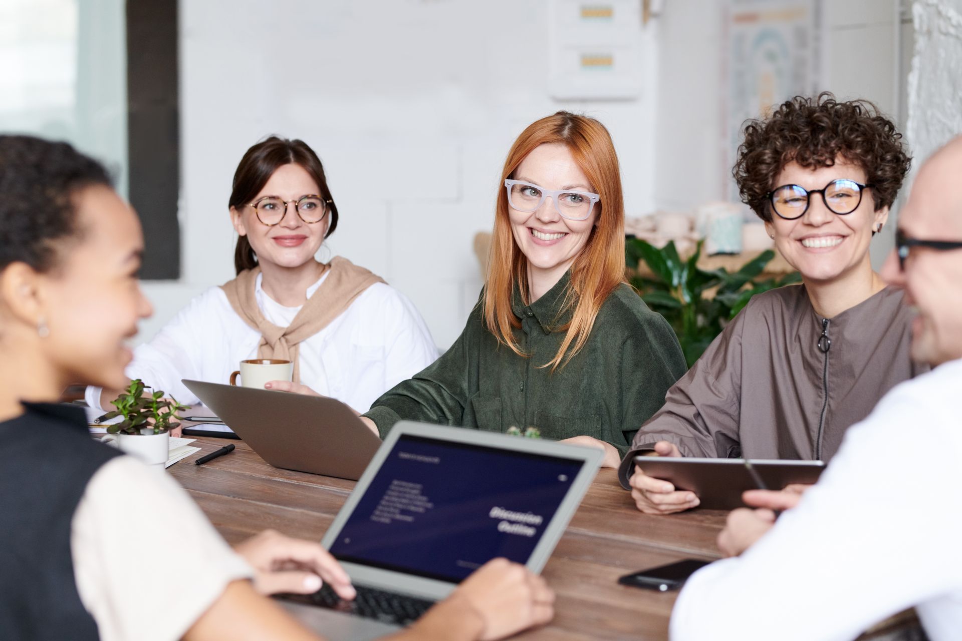 A woman with red hair and light glasses smiling at the camera between colleagues at a wooden meeting table