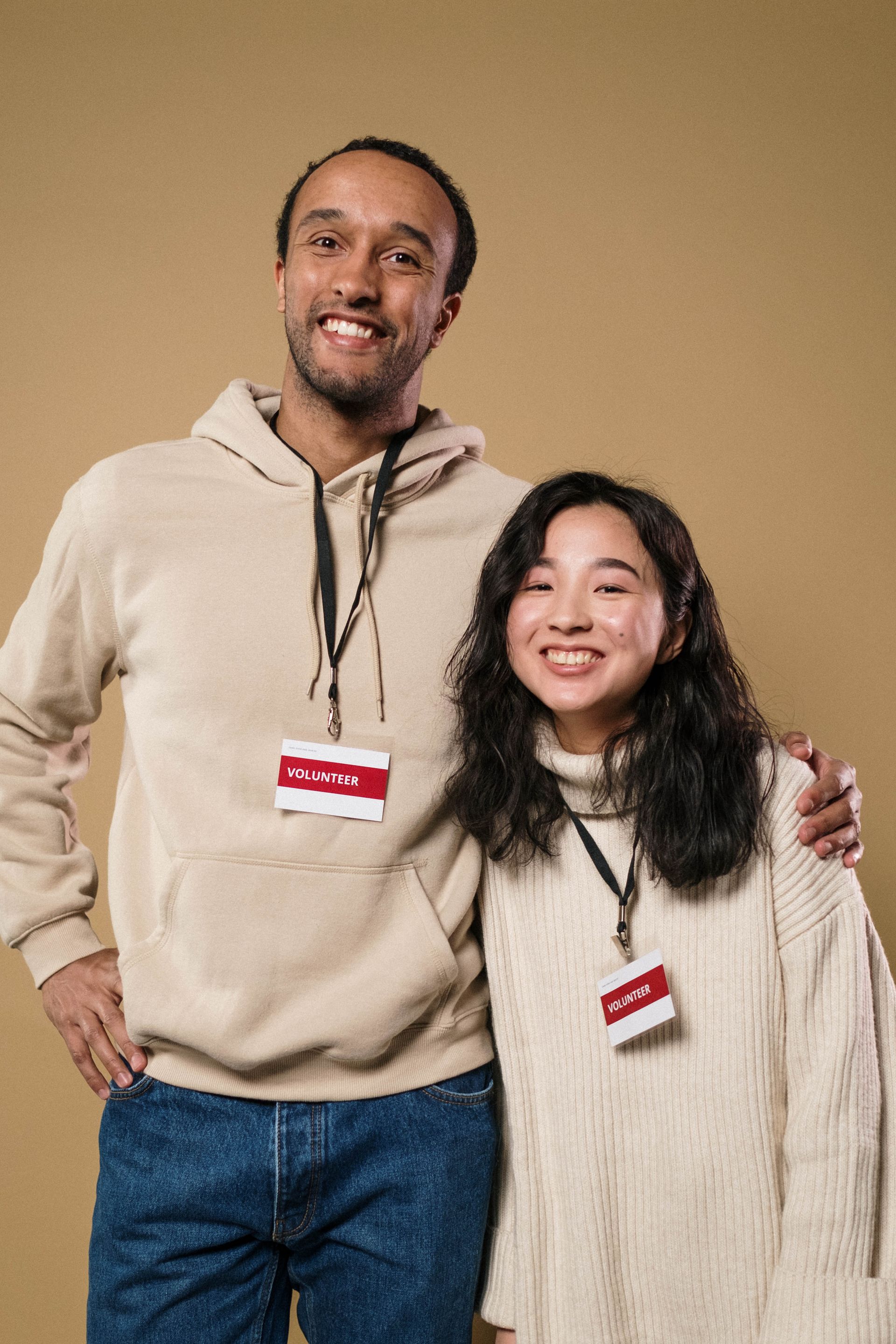 Two smiling volunteers in cream sweaters, wearing name tags, standing arm in arm against a warm tan backdrop