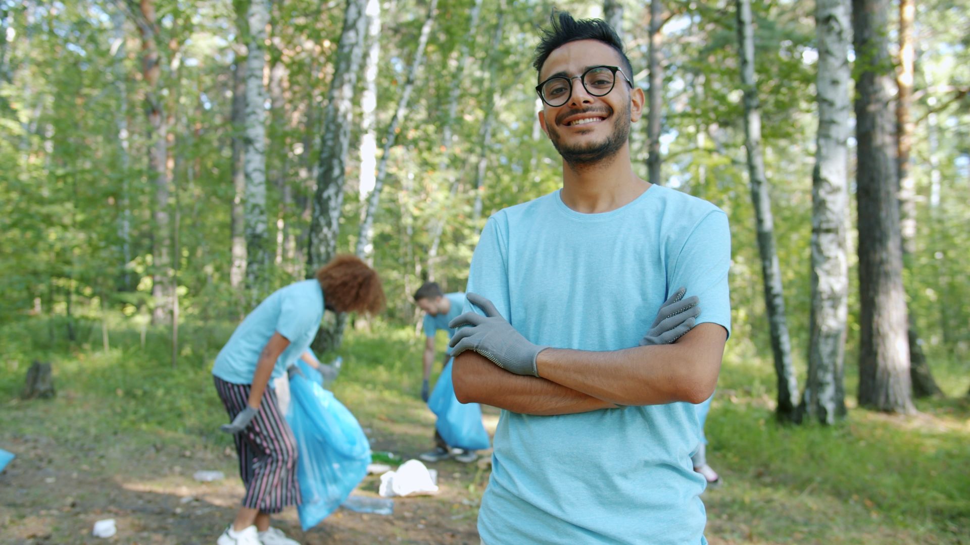 A smiling volunteer in glasses and work gloves standing in a sunny forest during a clean-up