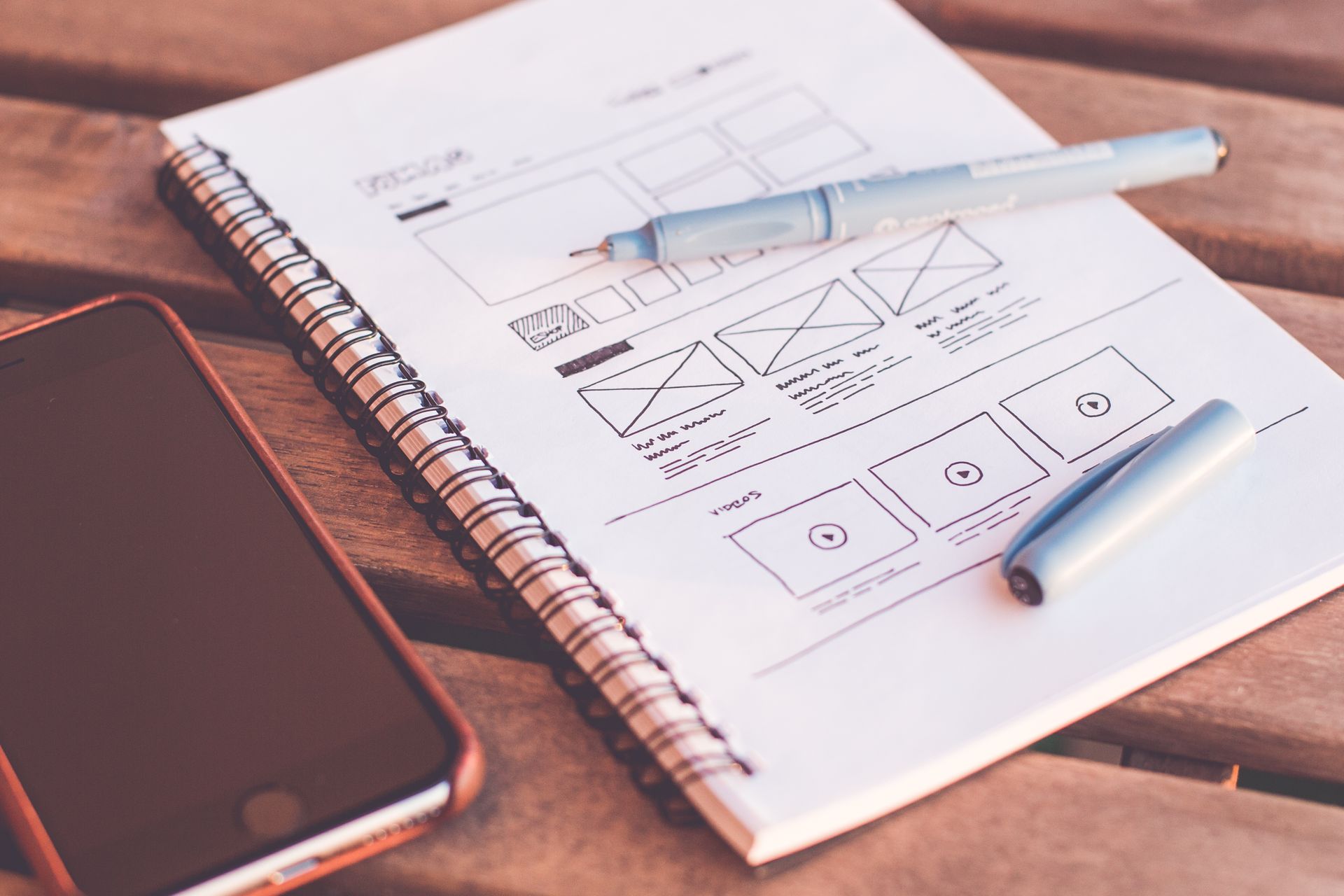 A notebook page with a hand-drawn website wireframe, a marker and a phone on a wooden table