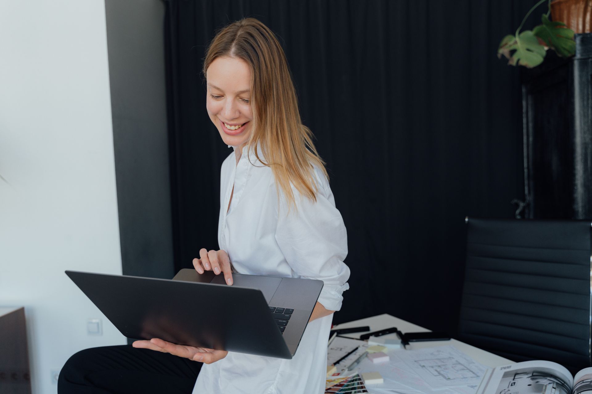 A smiling designer holding an open laptop beside a desk of plans and colour swatches